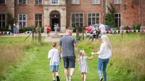 A family of four hold hands and walk through grassland towards Melford Hall while smiling.